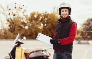 Courier Man In Uniform Offering To Sign Paper Holding Folder And Pen Standing Near Scooter Outside, Smiling To Camera photo