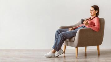A young woman with brunette hair is sitting comfortably in a light gray armchair, holding a cup. She is enjoying a moment of relaxation in a minimalist, bright setting. photo