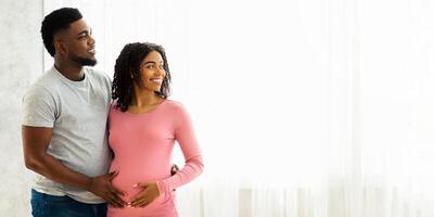 Happy african american pregnant family standing by window and looking at copy space for advertisement. Smiling black husband and expecting wife hugging at home, looking at advert for pregnant couples photo