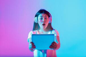 A young Asian woman with headphones looks amazed while holding a tablet in front of her. The background features a vibrant mix of pink and blue colors, creating a lively atmosphere. photo