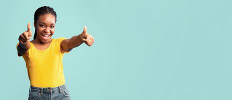 A young woman stands confidently, smiling and showing thumbs up with both hands. She wears a bright yellow top and high-waisted jeans, creating a lively and cheerful atmosphere. photo