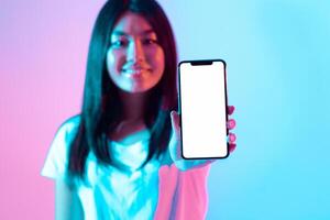 A young woman holds up a smartphone in front of her with a blank screen. The vibrant blue and pink background adds a modern touch to her confident pose in a studio setting. photo