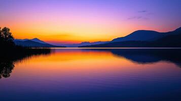 Vibrant hues of orange, purple, and blue fill the sky as the sun sets over a calm lake, surrounded by majestic mountains. The peaceful water reflects the stunning colors, creating a serene atmosphere photo