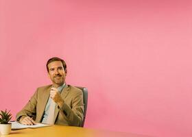 a man in a suit sitting at a desk with a pink wall behind him photo
