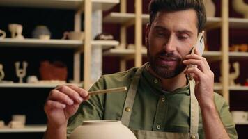 Talented Caucasian man male guy small business owner potter salesman businessman craftsman talking mobile phone call smartphone holding tool conversation making vase pottery ceramic order in workshop video