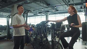 Personal trainer is communicating with an athlete who is cycling on an exercise bike in gym. man is explaining how to perform exercises correctly. video