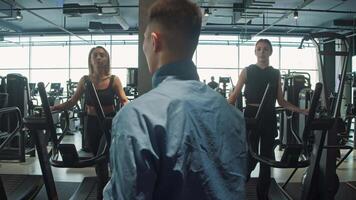 Two young female friends are exercising on a treadmill in gym, with the guidance of a fitness coach who explains the correct exercise techniques to them. video