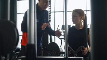 A young coach demonstrates and explains how to correctly perform exercises on a sports simulator to athlete training in the gym. A fragile girl lifts heavy weights in the gym video