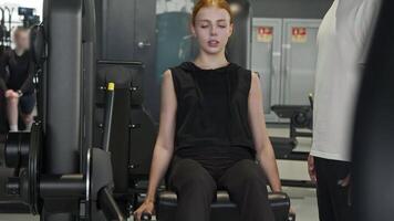 A young, attractive red-haired woman, under the guidance of a personal trainer, is doing exercises on a leg machine in the gym. video