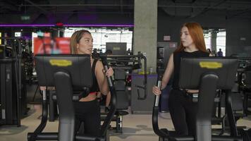 Two attractive young women in leggings and tops are chatting while working out on elliptical machines at the gym. video