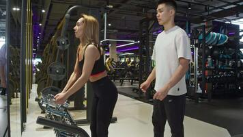 Personal trainer supports young woman in gym, providing guidance through series of exercises using dumbbells, which involve lifting with hands. video