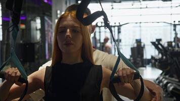 Young and attractive red-haired woman is seen pulling herself up on rings with assistance of her personal trainer. trainer helps her to perform exercise with correct technique in gym. video