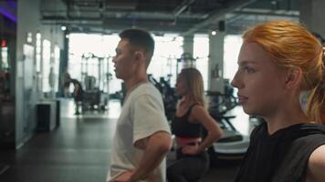 Young, attractive woman with red hair is warming up and training in front of the mirror, as part of group of athletes, before training at gym. video