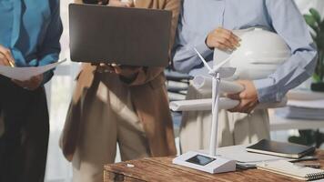 Three people standing around a table with laptops and a wind turbine video