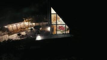 Night view of illuminated modern cabin with large windows, snowy yard and outdoor seating area. video