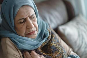 An elderly Middle-Eastern woman in a teal hijab expresses a moment of contemplation while sitting on a couch. photo