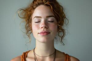 A young Caucasian woman with light curly hair and freckles, eyes closed in serene meditation against a soft gray background. photo