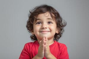 A joyful young boy with curly hair wearing a red shirt, clasping his hands together in excitement against a gray background. photo