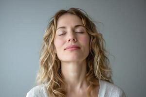 A serene young Caucasian woman with curly hair, eyes closed, reflecting tranquility against a soft gray background. photo