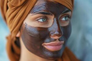 Close-up of a young woman with a chocolate facial mask, wearing a brown headwrap, showcasing her clear blue eyes and serene expression. photo