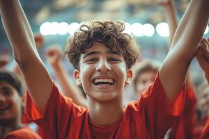 A joyful Middle-Eastern teenage boy celebrates in a vibrant red jersey, surrounded by fellow fans in a stadium filled with excitement. photo