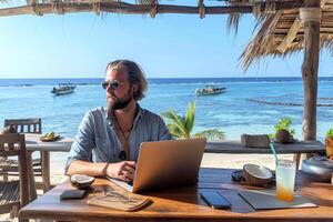A relaxed Caucasian man works remotely on a laptop at a tropical beach, enjoying the sea view with a refreshing drink by his side. photo
