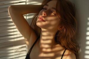 A serene young woman with wavy brown hair poses against a sunlit wall, shadows cascading over her peaceful expression. photo
