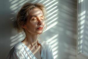 A serene Caucasian woman with closed eyes, illuminated by soft light casting shadows from a window, conveying peace and tranquility. photo