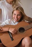 Portrait of a blonde girl playing guitar while smiling on camera with a woman sitting with he photo