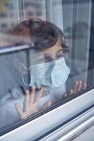 Pensive boy in mask standing alone and looking out of window at photo