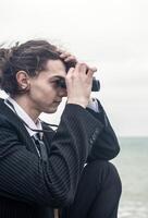 Portrait of a young man against the backdrop of the sea, he is looking into the distance through binoculars. He has long hair, is wearing a suit and tie. Relaxed, peaceful, thoughtful, happy and free on the seashore. photo