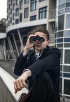 Portrait of a young man against the backdrop of modern architecture, he is looking through binoculars. He has long hair, he is wearing a suit. Front view, close-up. Relaxed, peaceful, thoughtful, happy photo