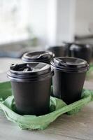 Three black plastic glasses with a drink stand on a light wooden table in the office. The glasses are covered with lids in a stand for transportation.. A cup of coffee on a table. photo