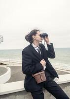A young man sits alone against the backdrop of modern architecture near the sea. He looks through binoculars at the sea. He has long hair, wears a suit and tie. Relaxed, peaceful, thoughtful photo