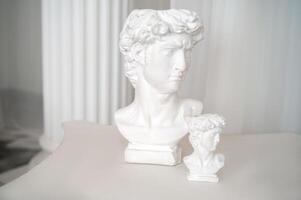 close up of beautiful antique decorative sculptures in interior. two plaster sculptures stand on antique style table. white chiffon fabric and columns behind photo