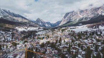 Aerial view of Cortina d'Ampezzo, showing snow covered rooftops, a yellow crane, preparing for the Winter Olympics 2026, dense forests, and dramatic Dolomite mountain peaks. video