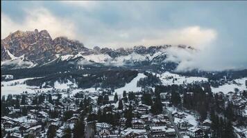 View of Cortina d'Ampezzo, Italy, with snow covered rooftops and winding roads. Clouds move over the Dolomite Mountains, casting light and shadow. video