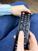 The TV remote control in hands of girl lies on her legs while watching an interesting television program. Control your TV at home using remote controller, vertical photo of TV controller
