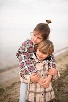 The brother and sister embrace and laugh on a wooden platform near the water, the cloudy sky setting a cozy mood. photo