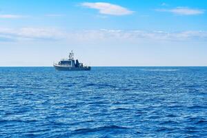 The boat is in the center of the photo, and the water is a deep blue color. The sky is clear and blue with white clouds on the horizon, and the waves are small and gentle. photo