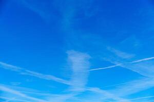 A beautiful shot of a light blue sky adorned with white wispy cirrus clouds. A few jet trails criss-cross the sky, creating an intricate pattern against the serene backdrop of nature. photo