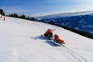 A snowmobile rescuer is driving a rescue sled with an injured skier on it. The background is a beautiful mountain landscape with snow-covered trees and mountains. photo