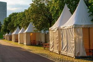 The image features white party tents arranged neatly in a park, with a vibrant green landscape and trees providing a beautiful backdrop. The tents have peaked roofs and line the pathway. photo