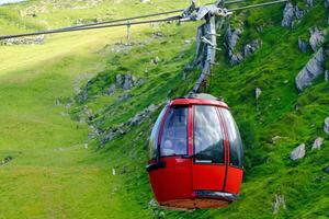 A red cable car cabin with glass windows travels over a green mountainside. The cabin is suspended from a cable, surrounded by trees and grass, captured from a low angle. photo
