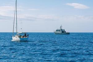 A small sailboat with three individuals is being inspected by an armed forces vessel in the open sea, showcasing maritime security operations and the importance of safeguarding waters. photo
