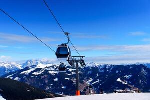 This is an isolated image of a modern ski lift gondola in the Alps. The gondola is white and has a black trim. It is surrounded by snow-capped mountains and a blue sky. photo
