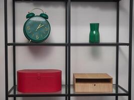 A green alarm clock sits on a shelf next to a red box and a green vase photo