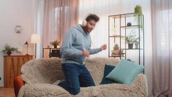 Young man dancing on couch with wireless headphones playing air guitar and enjoying disco music video