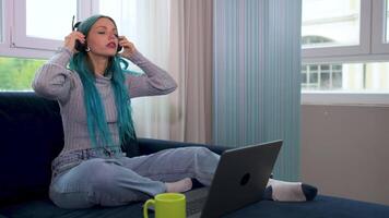 A beautiful blue-haired hipster woman is working from home sitting on her couch, there is a problem, annoyed she puts on headphones with microphone to participate in a meeting with her laptop. video
