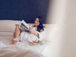 Smiling woman reading magazine during breakfast on bed photo
